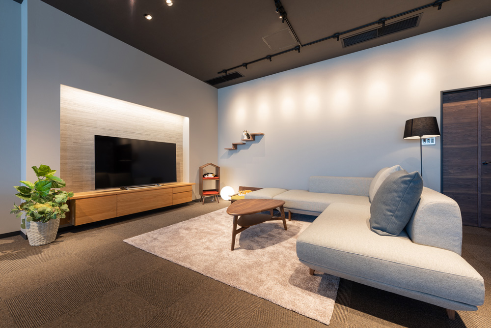 広々としたリビングルームに、大型テレビと木製のテレビ台、L字型のグレーのソファ、丸いコーヒーテーブルが配置されています。観葉植物と温かみのある照明が心地よい雰囲気を演出しています。