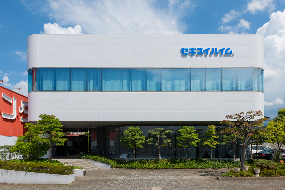 セキスイハイム東北山形支店の社屋写真。白い壁と大きなガラス窓が特徴で、建物の前には緑の木々が並んでいます。青い空に建物のロゴが映えています。