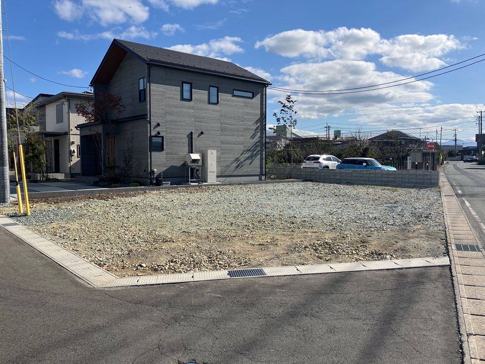 欅タウン分譲地の写真です。角地に広がる砂利が敷かれた分譲地で、隣には灰色の二階建ての住宅が建っています。晴れた青空の下、白い雲がぽっかり浮かび、穏やかな住宅街の一角が広がっています。