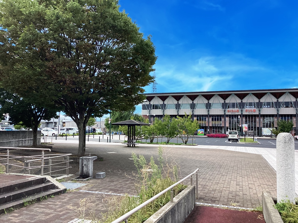 さくらんぼ公園、さくらんぼ東根駅の写真。大きな木が茂る広場の向こうに、ガラス張りの大きな建物が見えます。建物前には駐車場があり、少数の車が停まっています。青空が広がり、穏やかな日差しが感じられる、落ち着いた街の風景です。