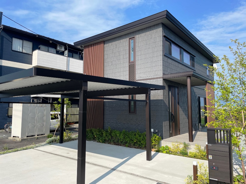 ブラックの外壁の二階建て住宅の外観。黒い外壁に縦の木目調の装飾が施され、前にはカーポートと植栽された庭が広がっています。新築らしい清潔感が漂っています。