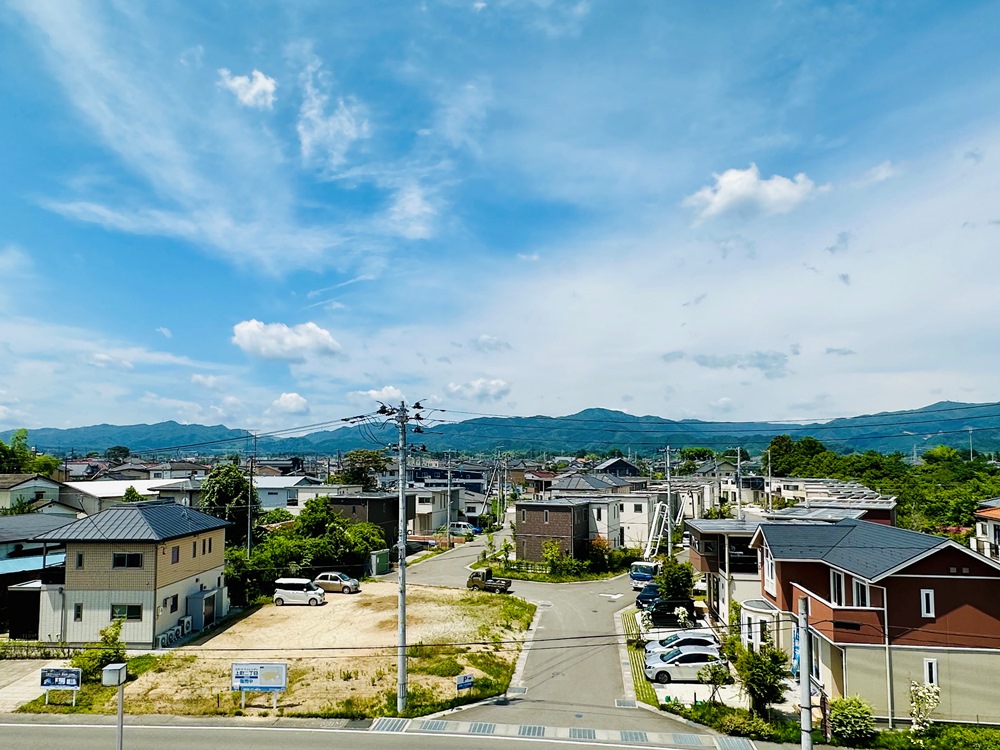 青空の下にスマートハイムシティ上町二丁目が広がり、遠くに緑の山々が見えます。家々の間には電柱や電線があり、住宅地の一部は空き地です。穏やかな午後の光が町全体を照らしており、静かで落ち着いた雰囲気です。