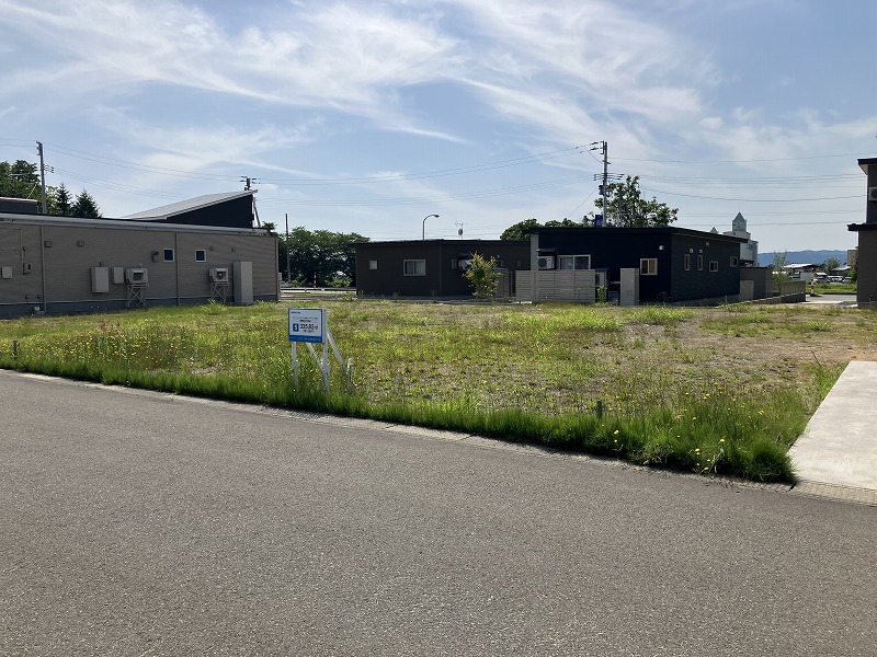 広大な空き地の写真で、中央に販売区画の看板があり、背景に家が二軒建っています。