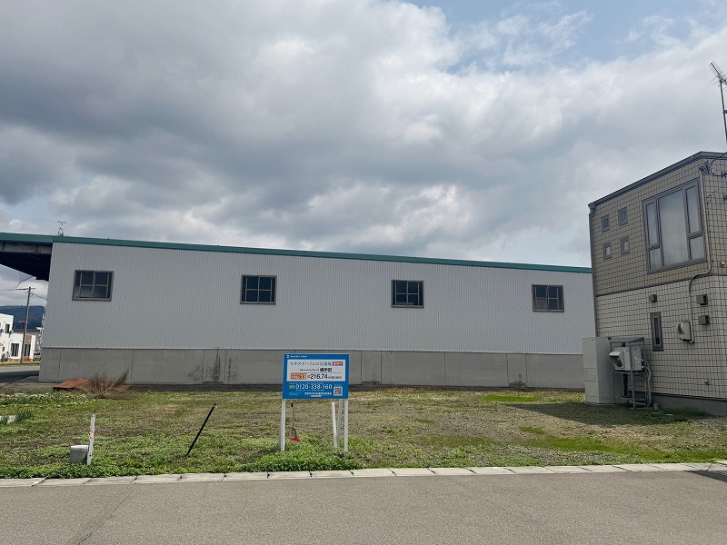 分譲地の右手にはグレーの建物、奥には白い外壁の大きな建物があります。手前には不動産案内の看板が立っています。