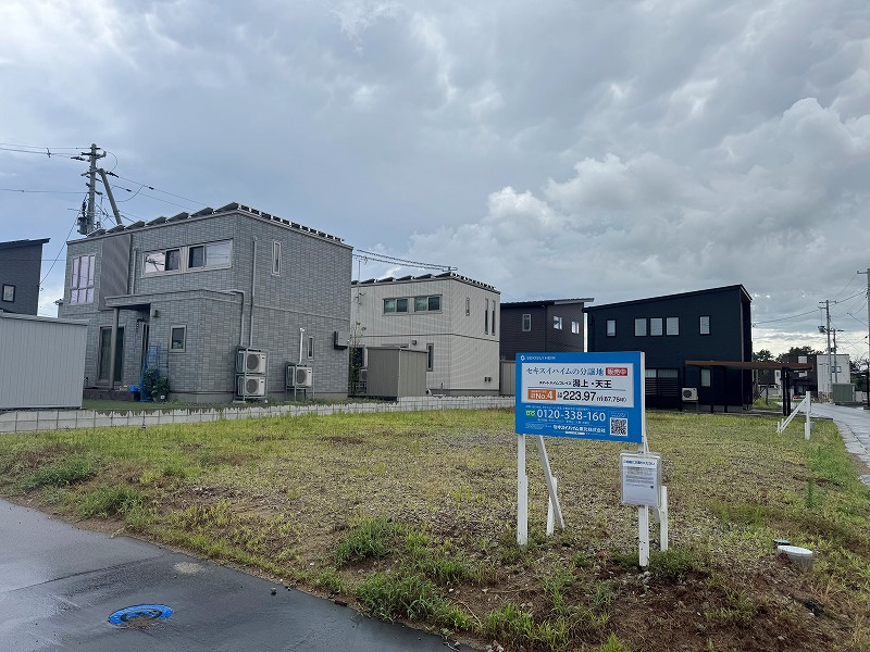 住宅地内にある分譲地。周囲には黒や白を基調としたセキスイハイムの住宅などが建ち並んでいます。道路沿いには電柱や駐車された車が見えます。