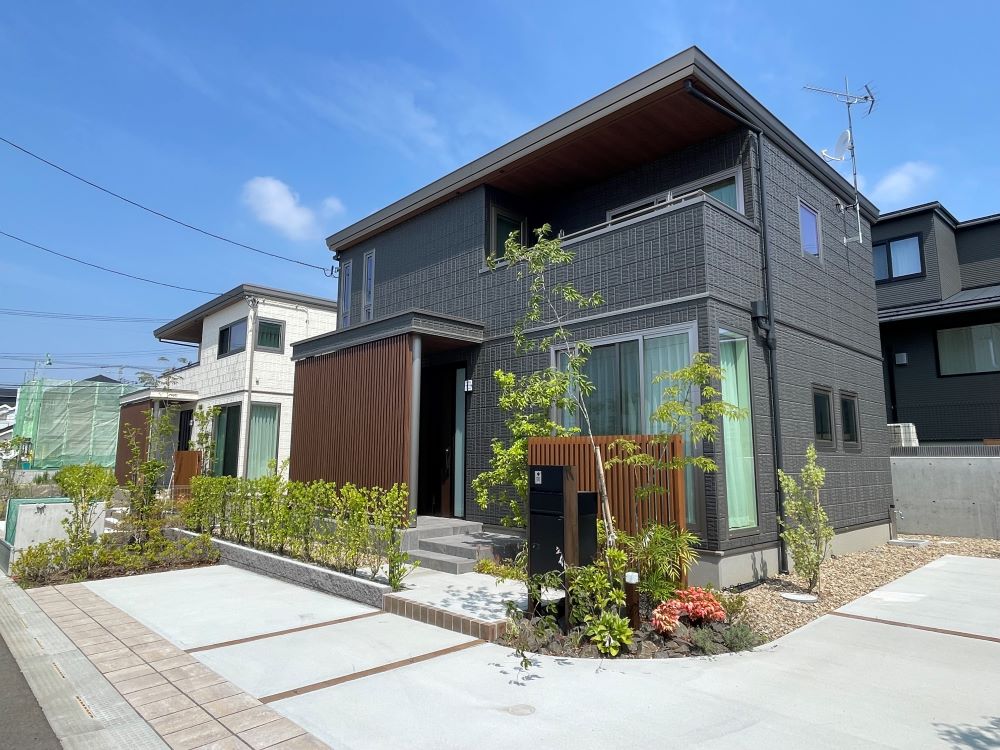 ダークグレーの外壁の２階建て住宅の外観。と奥には白い外壁の２階建て住宅が見えます。植栽が植えられています。
