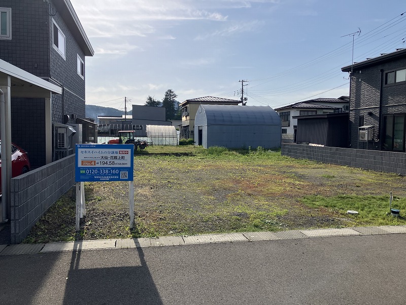 住宅街にある更地の分譲地。前面には分譲案内板が設置され、区画番号と面積、問い合わせ先などの情報が記載されている。周囲には戸建て住宅が並び、敷地奥には倉庫やビニールハウスのような建物が見える。