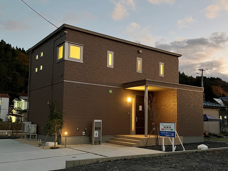 グレーを基調とした二階建て住宅の外観。縦長の窓が特徴で、左側には玄関への庇が設けられています。バルコニーにはアンテナが設置され、敷地内には小さな植栽が並んでいます。
