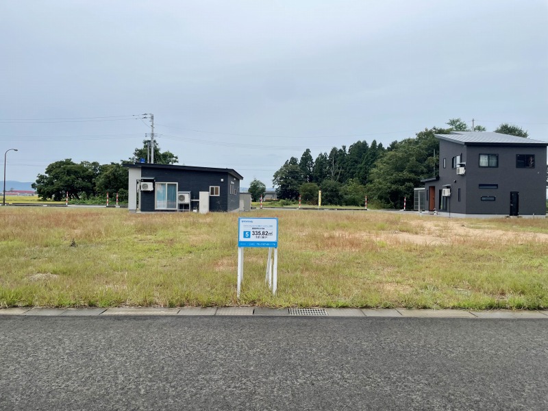 広大な空き地の写真で、中央に販売区画の看板があり、背景に家が二軒建っています。