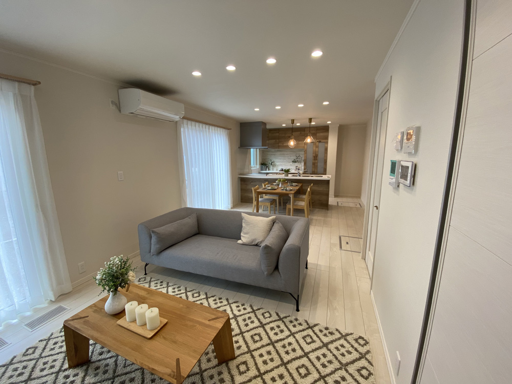 リビングルームの写真です。灰色のソファが中央に置かれ、木製のローテーブルの上にはキャンドルが並べられています。大きな窓には白いカーテンがかかり、自然光が優しく差し込んでいます。奥には、木目調のキッチンが見え、天井からは2つのペンダントライトが吊るされています。