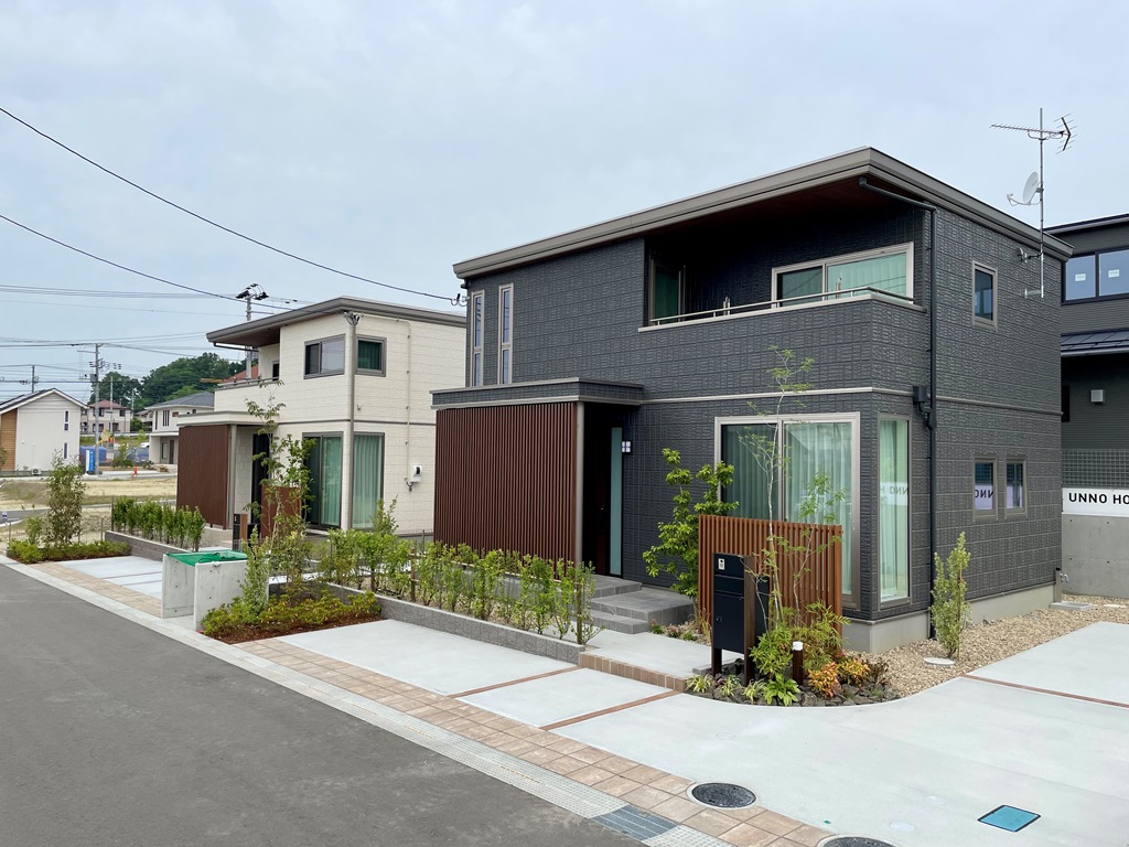 ダークグレーの外壁の２階建て住宅の外観。と奥には白い外壁の２階建て住宅が見えます。植栽が植えられています。