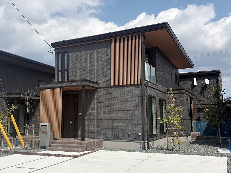 黒い外壁と木目調のアクセントが特徴的な二階建ての住宅です。正面には玄関があり、階段を上がると入り口に続きます。敷地内には小さな植木も植えられ、落ち着いた雰囲気を感じさせます。