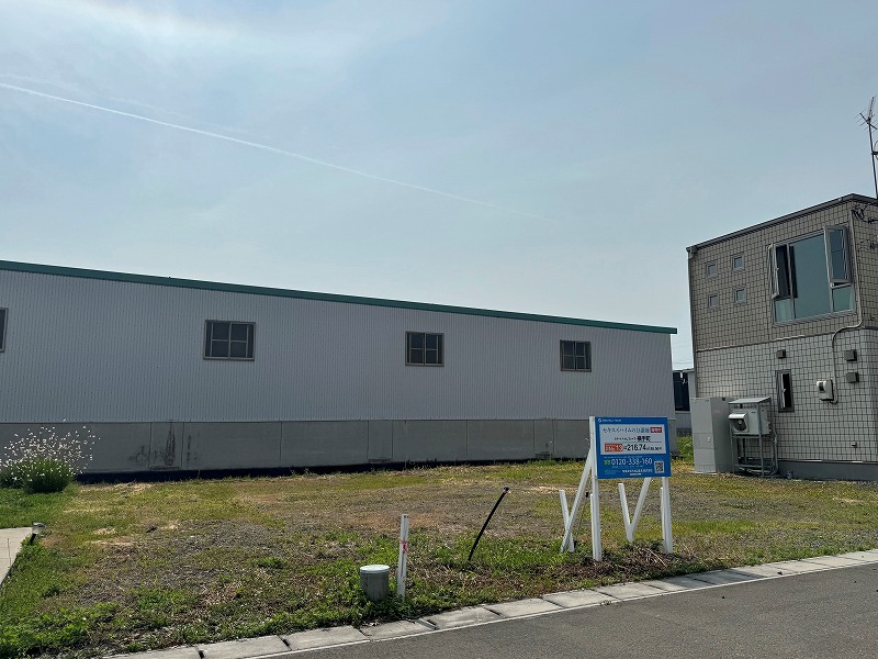 分譲地の右手にはグレーの建物、奥には白い外壁の大きな建物があります。手前には不動産案内の看板が立っています。