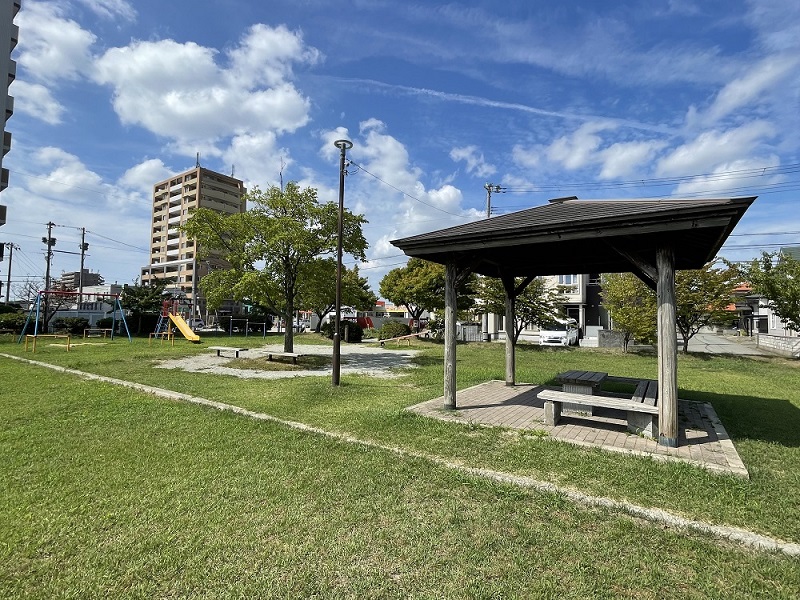 ベンチ付きの木の東屋と遊具が見える緑豊かな公園。左奥にマンションが見えます。