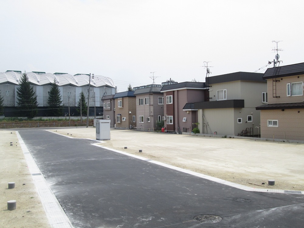 数軒の住宅が並ぶ分譲地で、前面道路は舗装されています。背景には大きな建物と木々が見え、静かな環境が広がっています。