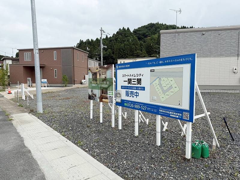 「スマートハイムシティ一関三関」と書かれた区画販売の案内板が立つ宅地。後方には住宅と緑の木々が見える静かな住宅地。