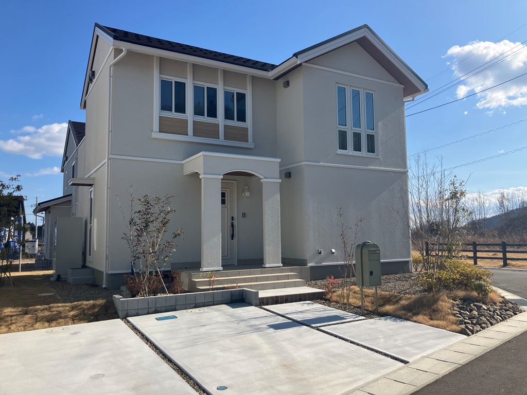 白い外壁の住宅が建っており、赤茶色の屋根が特徴です。シンプルで落ち着いたデザインで、前には駐車スペースと植栽が整備されています。