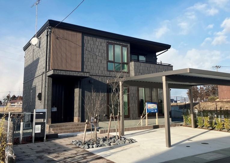 ダークグレーの外壁の二階建て住宅の外観。玄関前には小さな木が植えられ、右手にはカーポートがあります。周囲には緑の緑が広がり、広々とした景色が広がっています。