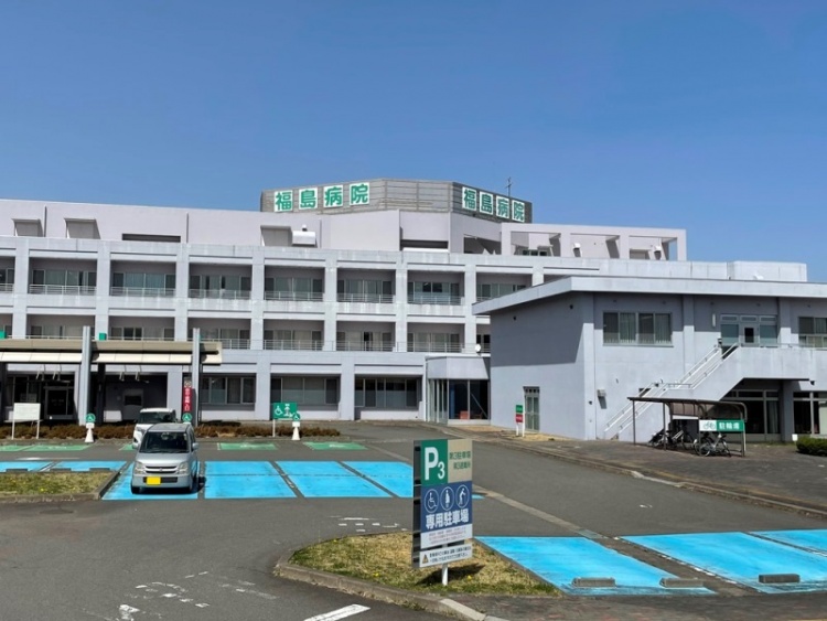 白い建物に緑の文字が特徴的な福島病院の外観。手前には駐車場が見えます。