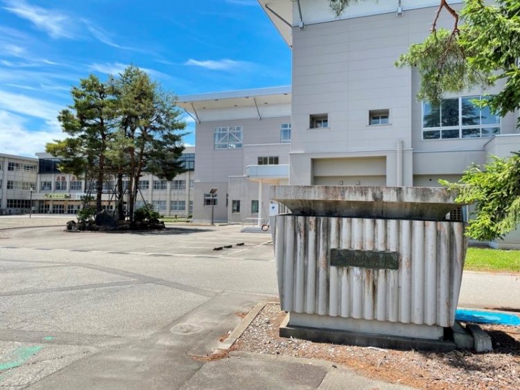 白い建物と植栽が見える大仙市立大曲中学校の校庭の風景。手前に学校の表札と樹木があります