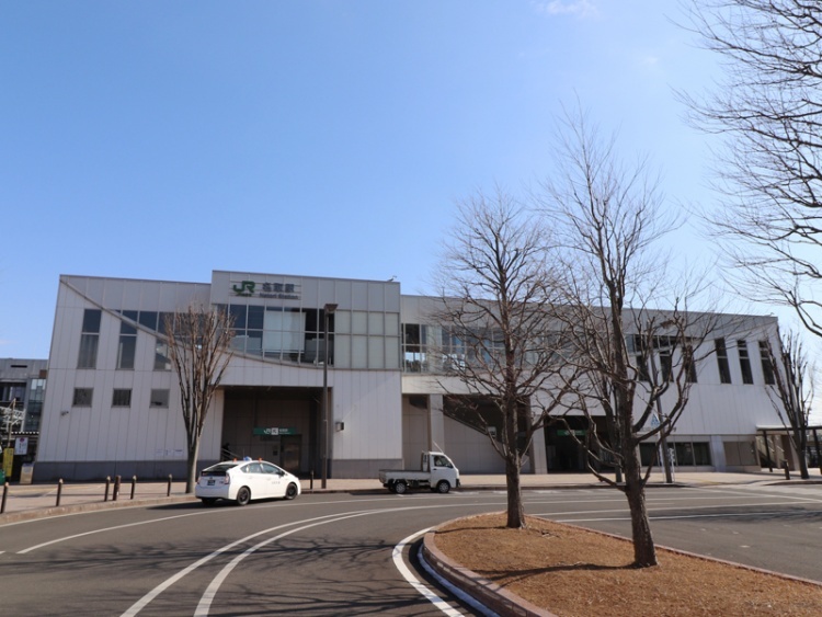 白い外壁のJR名取駅の駅舎。手前にロータリーがあり、周辺には木々が見えます。