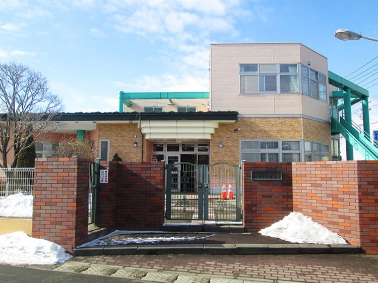 赤いレンガの門とフェンスに囲まれた二階建てのこざくら保育園の建物。周囲には雪が少し残っています。