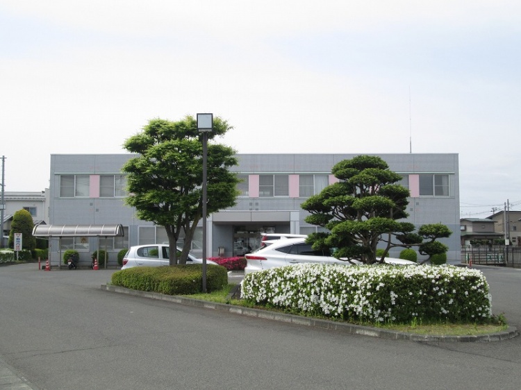 グレーとピンクの外壁が特徴的な三愛病院矢巾クリニックの外観。植栽が植えられていて、その奥に駐車場が見えます。