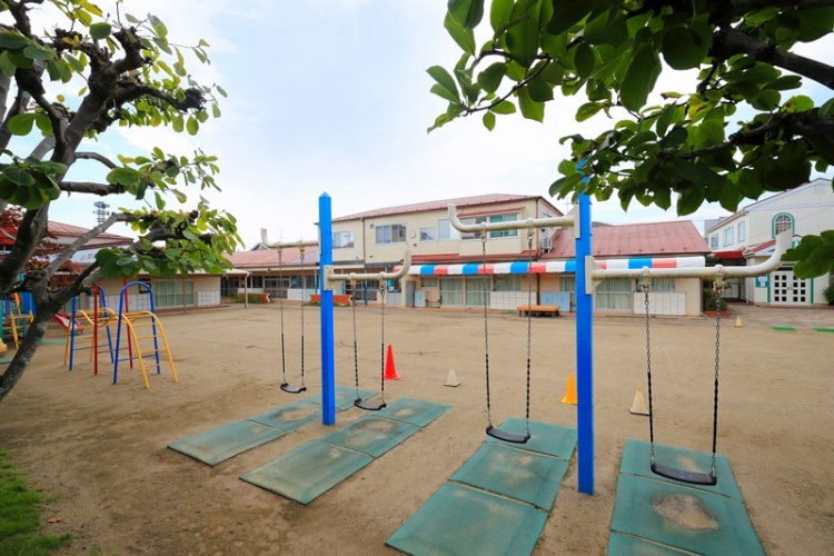 赤い屋根が特徴的なしげる幼稚園の外観。園庭にはブランコや遊具があり、奥には園舎が見えます。木々に囲まれています。
