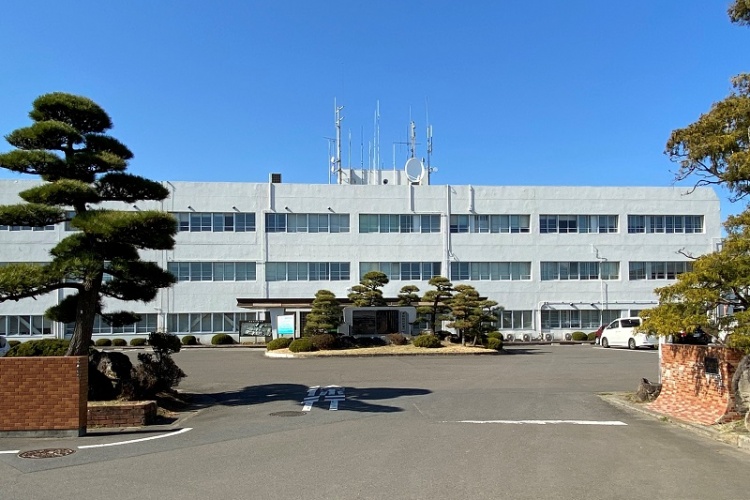 白い外壁の二階建ての東松島市役所の外観。前には整えられた松の木と舗装された駐車場が見えます。