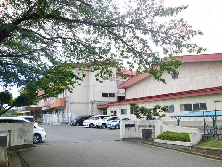 コンクリート造りが特徴的な高松小学校校舎の外観。駐車場には複数の車が停められています。周囲には木々が生えています。
