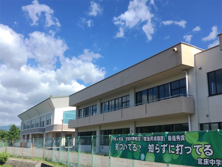 白とベージュの外壁の宮床中学校校舎の外観。緑のバナーが掲げられたフェンスがあります。