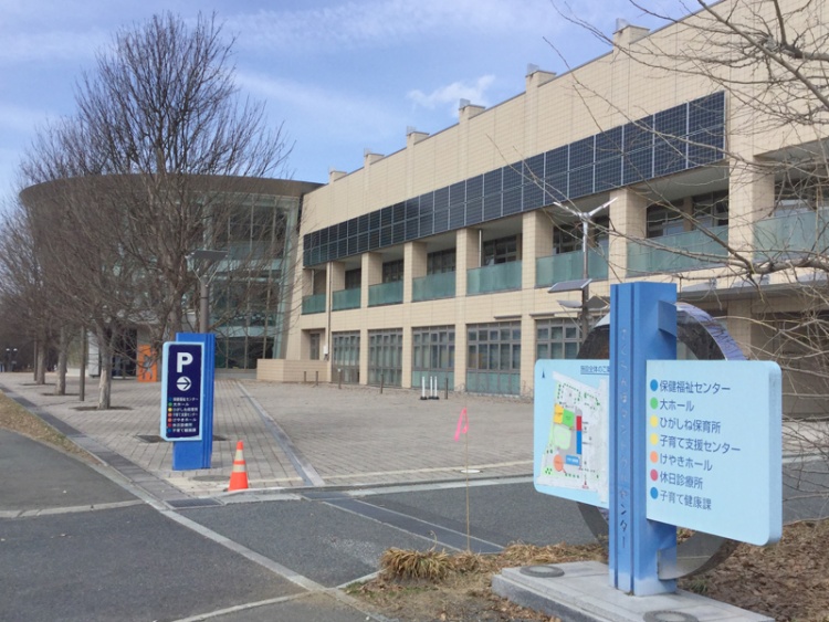 白い壁の大きな建物のサクランボタンクトクルセンターの外観。前に駐車場案内や施設案内の標識があります。歩道と自動車用の道が見えます。