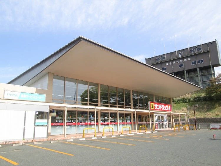 大きな屋根とガラス張りが特徴的なサンドラッグ三ツ割店。広い駐車場が見えます。