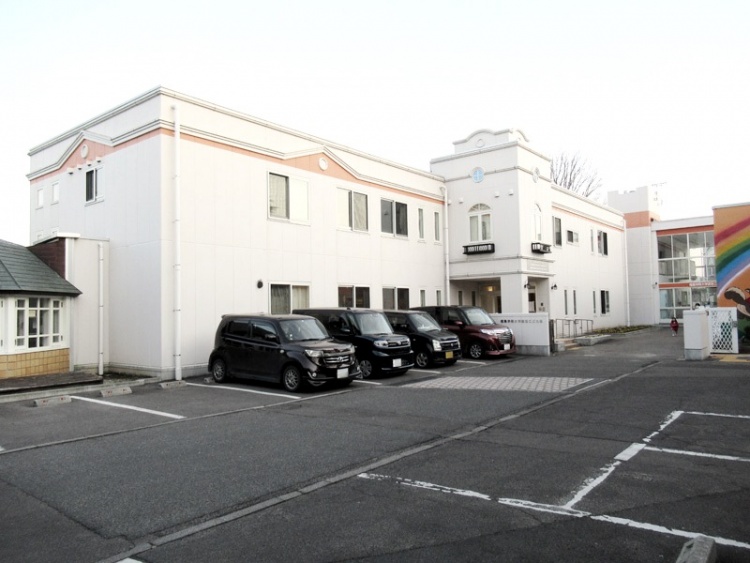 白とピンクの2階建ての建物で建物の右側には虹の絵が描かれて。駐車場には数台の車が停まっています。