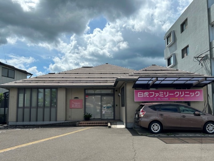 玄関とピンクの看板が特徴的な白虎ファミリークリニックの外観。駐車場には1台の車が停められています。