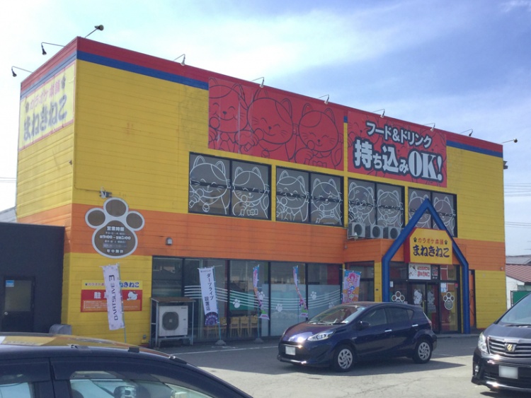 黄色の外壁が特徴的なカラオケ店まねきねこ東根店の外観。持ち込みOKの看板が書いてあります。駐車場スペースがあります。