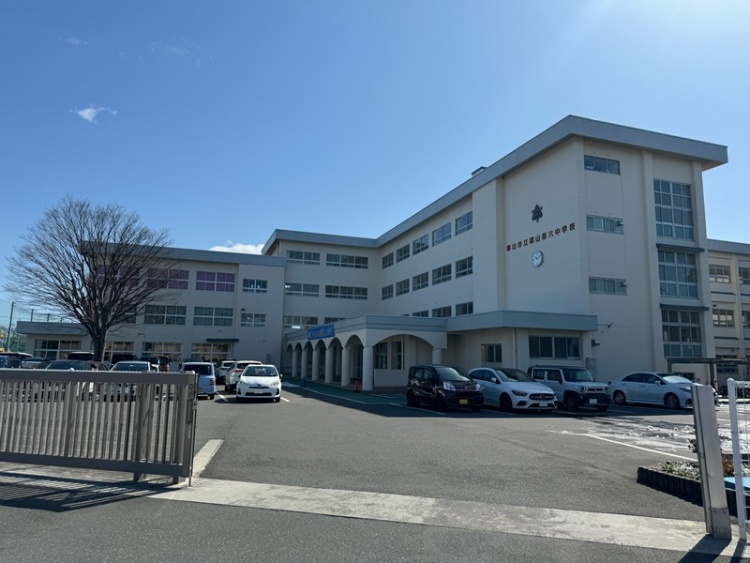 ベージュの外壁とグレーの屋根が特徴的な郡山第六中学校の校舎の外観。駐車場には車が数台停まっています。