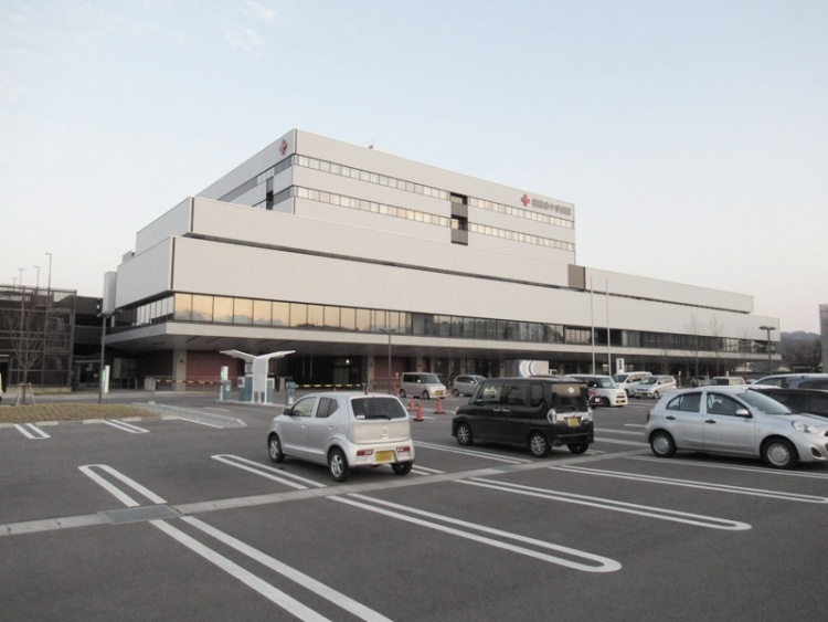 広い駐車場に隣接する大きな白い建物が特徴的な赤十字病院。駐車場には数台の車が駐車されています。