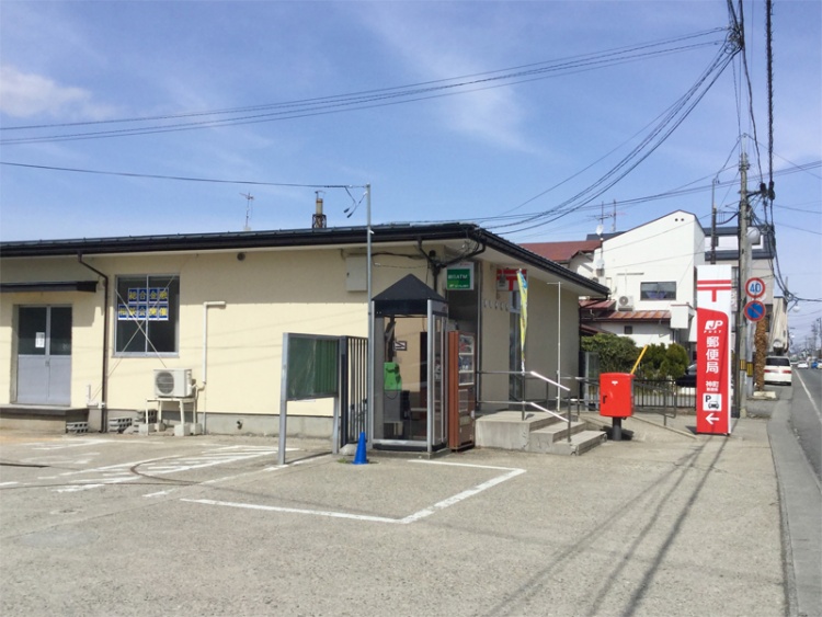 薄黄色の建物に神町郵便局の外観。正面には赤い郵便ポストと駐車場が見えます。