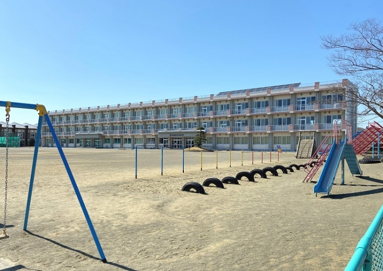 3階建ての矢本西小学校校舎の外観。校庭にはタイヤや鉄棒、滑り台等が設置されています。