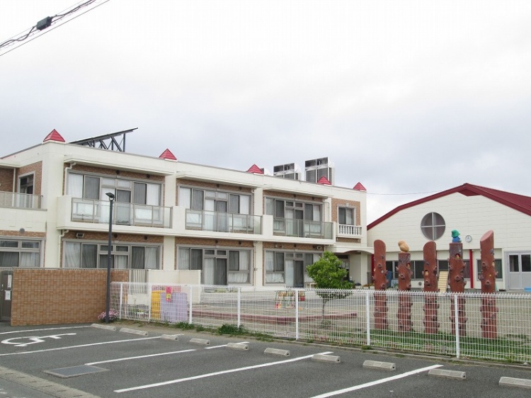 2階建ての建物と遊具がある幼稚園の外観。駐車場が手前にあり、フェンスで囲まれています。