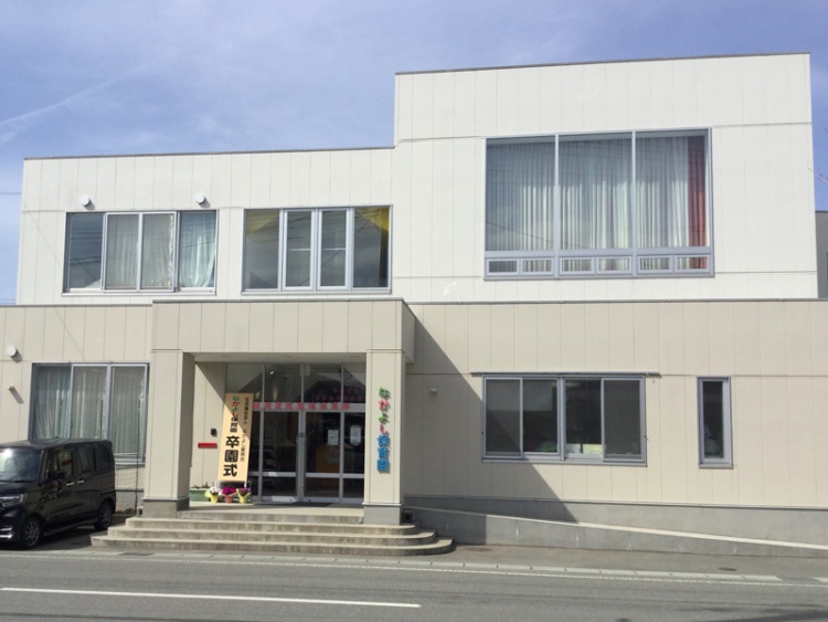 白い外壁の2階建てのなかよし保育園の外観。玄関横には卒園式の看板が立っています。
