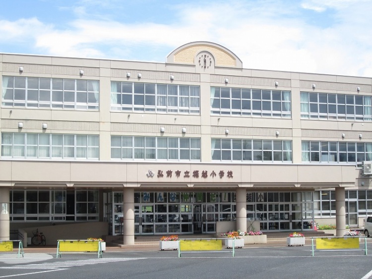 弘前市立堀越小学校の正面外観。3階建ての建物で、弘前市立堀越小学校の校名表示があります。正面玄関に花壇があり、植栽が見えます。