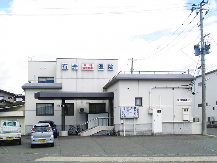 青い文字で書かれた石井内科消化器科の外観。建物前に駐車場があり、数台の車が停められています。