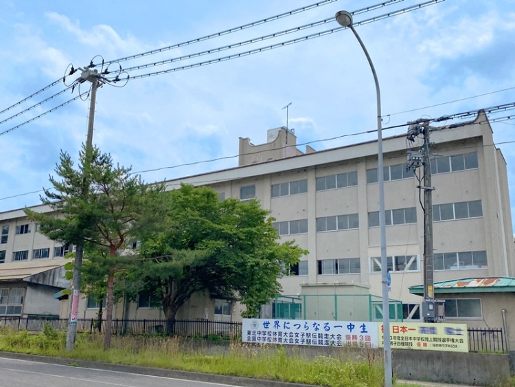 白い外壁の第一中学校校舎の外観。正面には世界に つらなる一中生と書かれた看板が見えます。