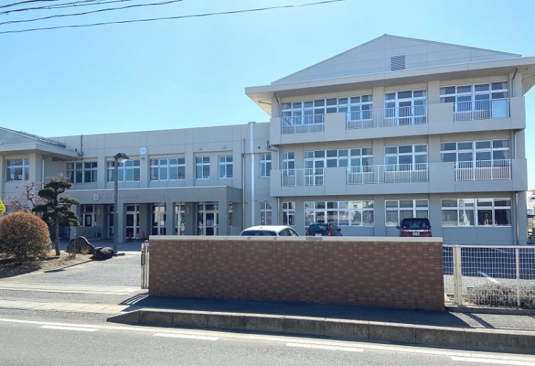 三角の屋根が特徴的な3階建ての矢本第一中学校校舎の外観。手前には駐車場と道路が見えます。