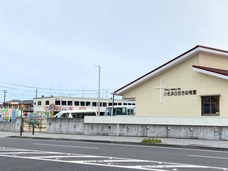 白い壁と赤い屋根が特徴的な小名浜白百合幼稚園の外観。園庭には遊具があり、バスが停まっています。