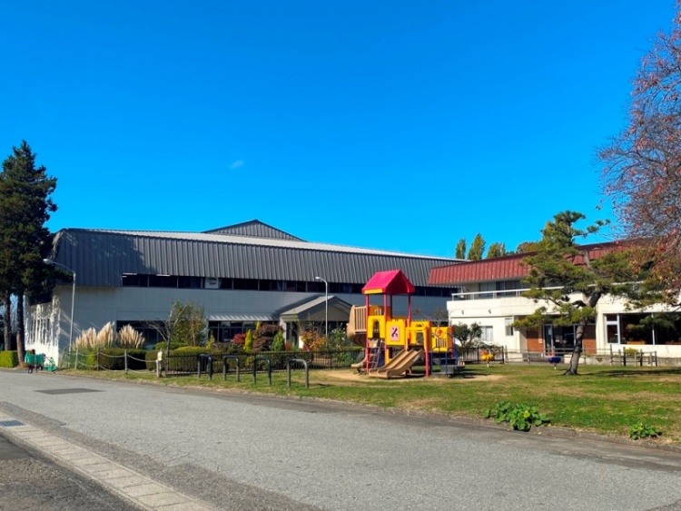 広場には赤と黄色の遊具があり、奥には市民ふれあいスポーツセンターの建物が見えます。