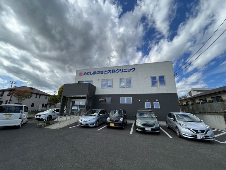 白とグレーの外壁の2階建てのめでしまのさと内科クリニックの外観。手前には駐車場が見えます。