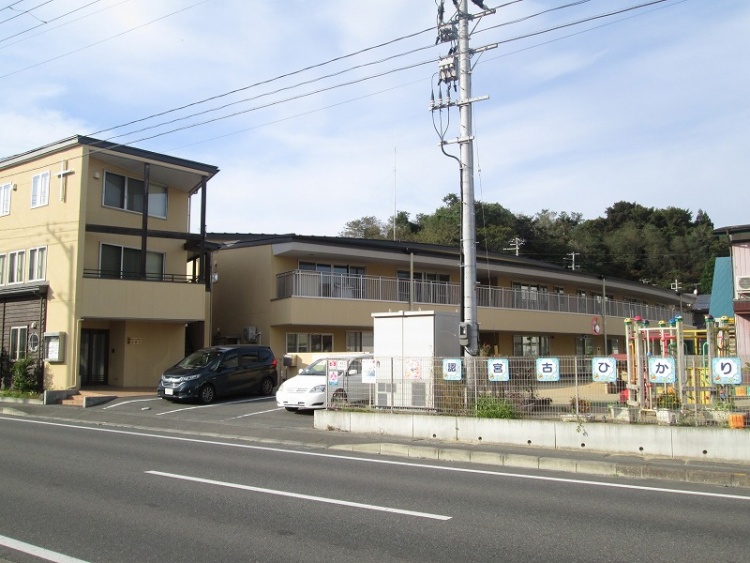 薄黄色の外壁が特徴的や宮古ひかりこども園の外観。フェンスの内側には遊具が見えます。駐車場もあります。